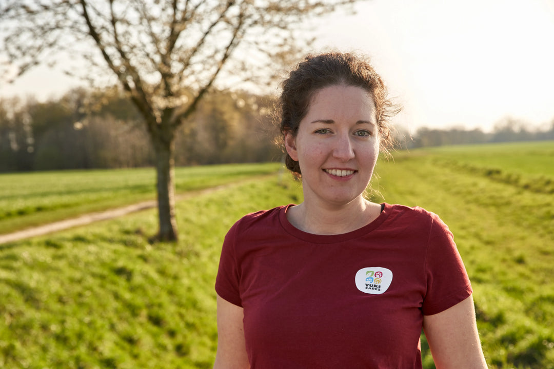 Eine Frau, Katrin, steht lächelnd auf einem grünen Feldweg, im Hintergrund ein Baum und ein sonniger Himmel.