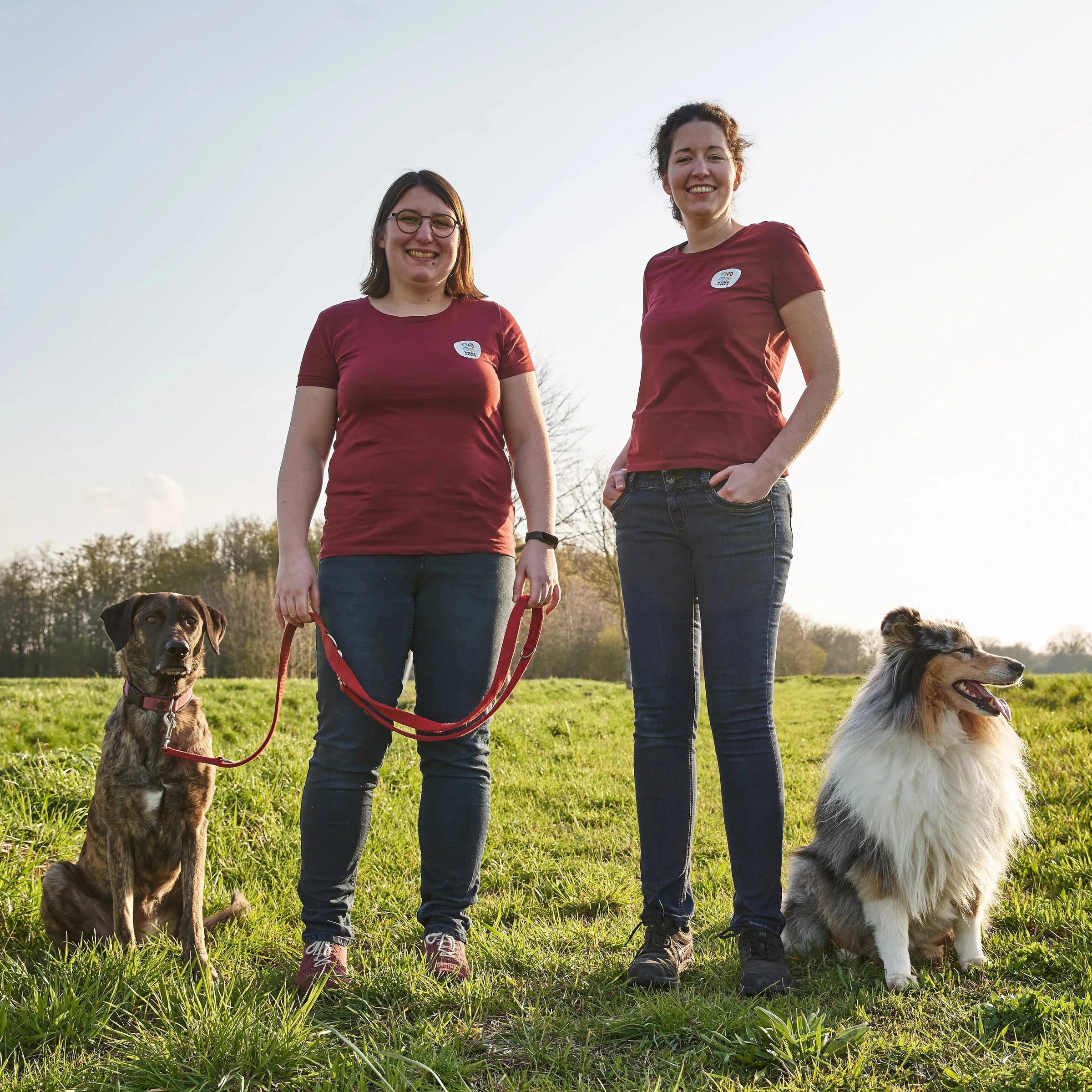 Christina und Katrin stehen mit ihren Hunden auf einer grünen Wiese und tragen passende rote T-Shirts unter blauem Himmel.