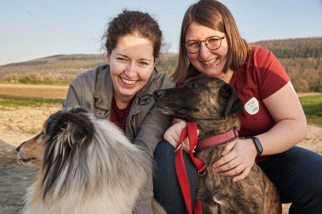 Zwei lächelnde Frauen posieren mit ihren Hunden im Freien, während die Sonne über der Landschaft untergeht.