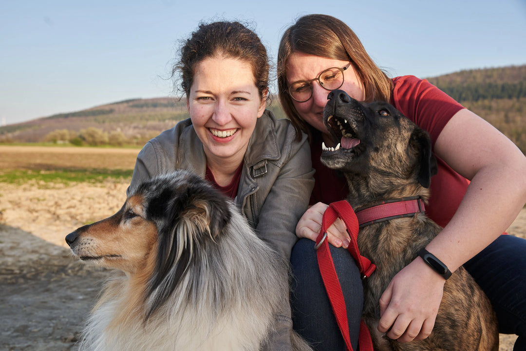 Zwei Frauen lachen glücklich in die Kamera, während sie mit ihren zwei Hunden in einer ländlichen Landschaft posieren.
