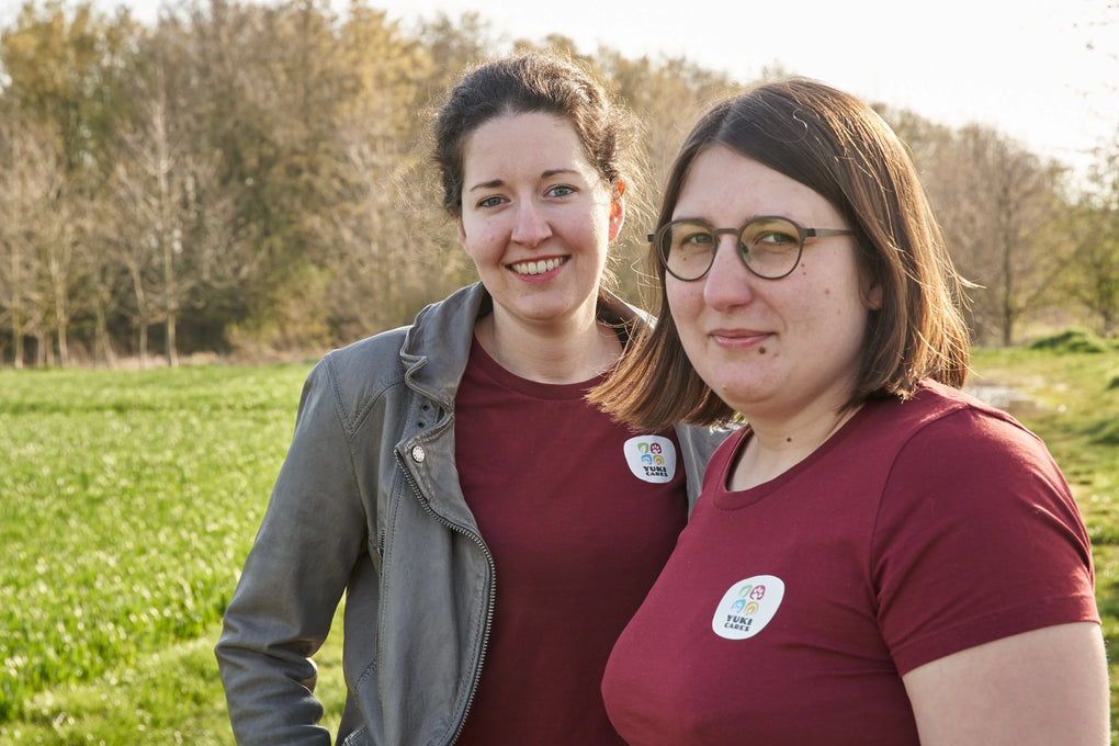 Christina und Katzen stehen lächelnd auf einem Feldweg, tragen rote T-Shirts mit "Yuki Cares"-Abzeichen und genießen den sonnigen Tag.