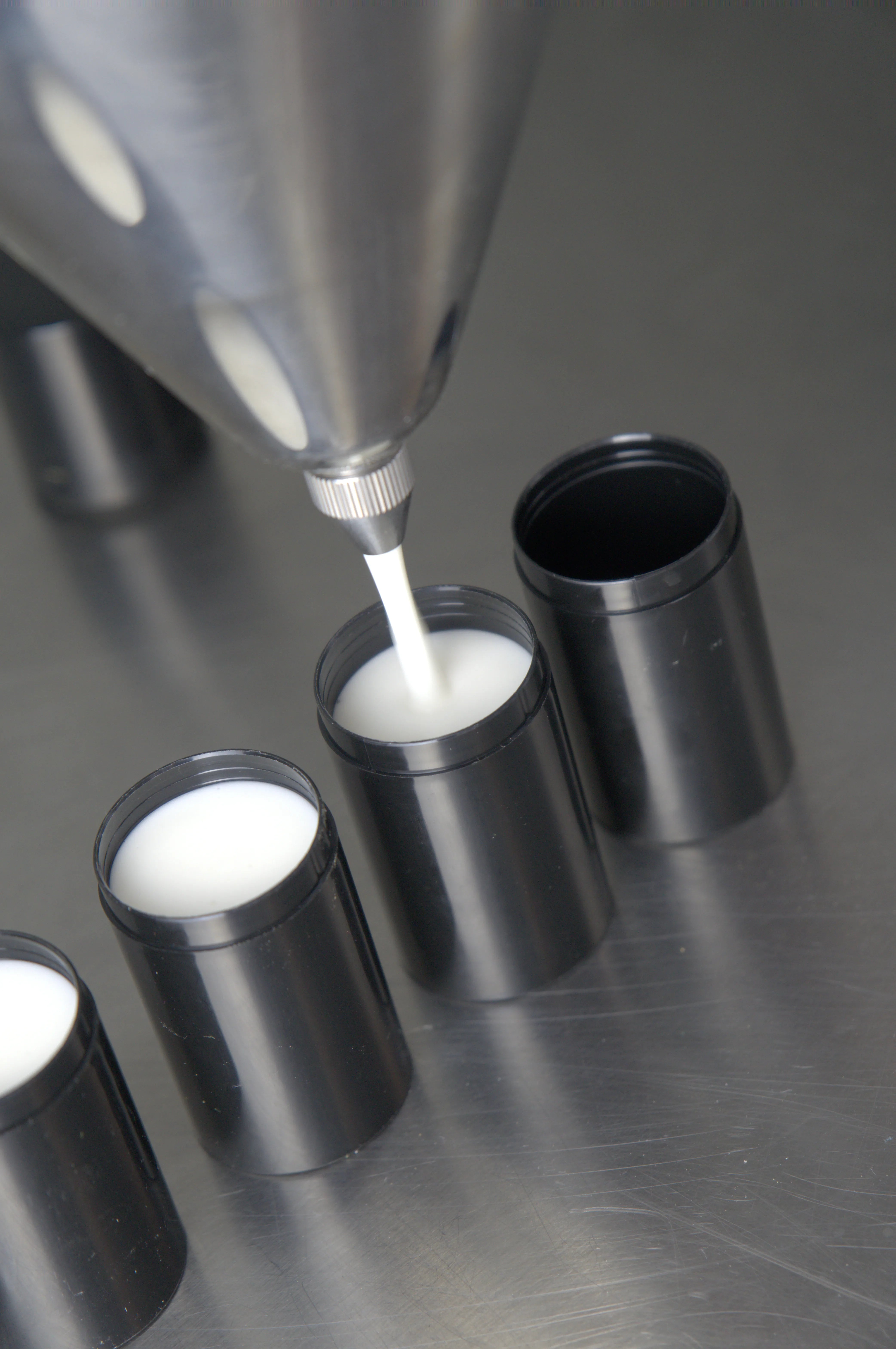 Eine Düse füllt weiße Creme in schwarze Behälter auf einer metallischen Oberfläche.