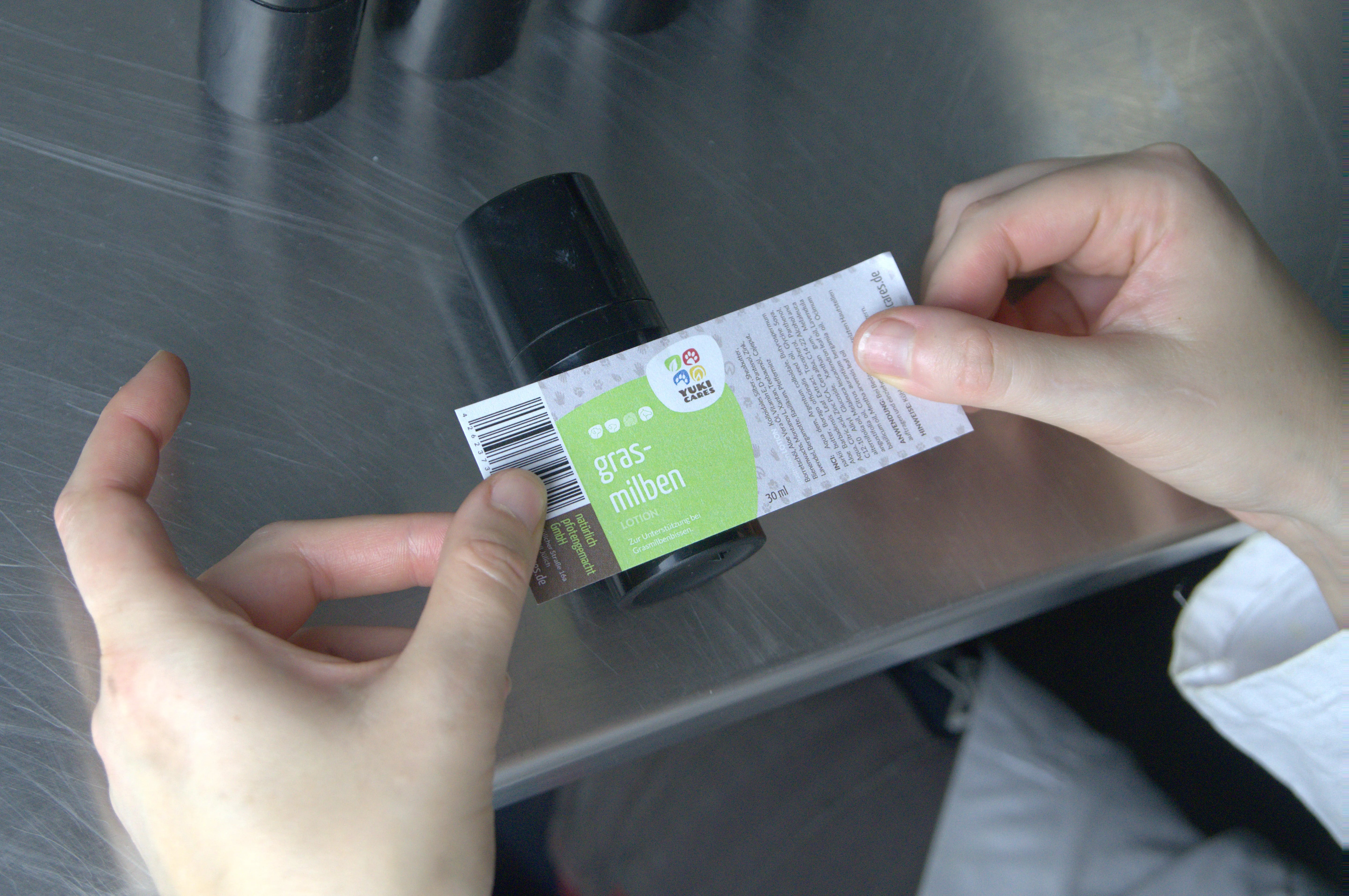 Zwei Hände kleben ein Etikett mit der Aufschrift "Grasmilben Lotion" auf eine kleine schwarze Flasche.