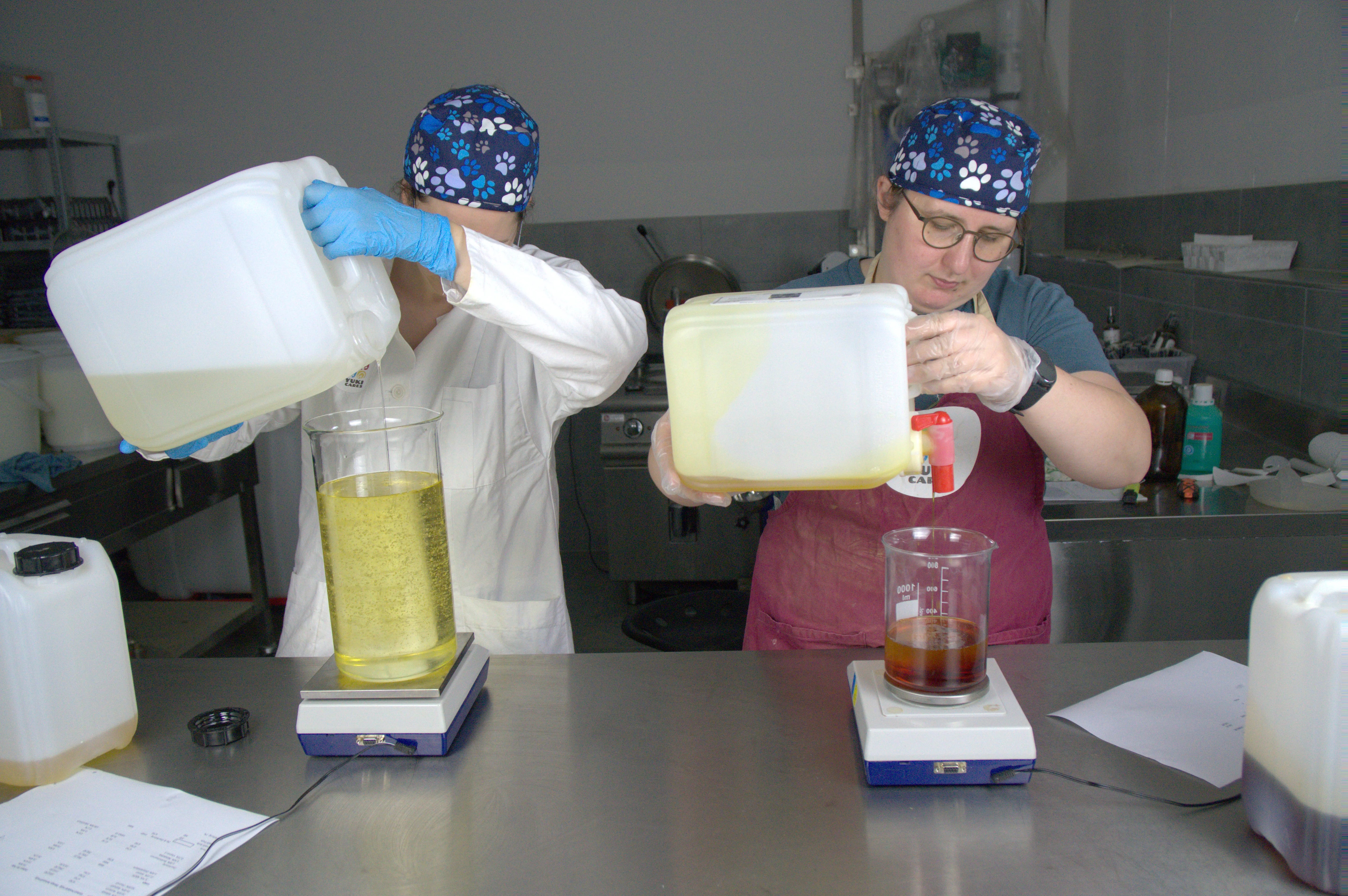 Zwei Personen in Hygienekleidung füllen Flüssigkeiten aus Kanistern in Messbecher auf einer Metalloberfläche ab.