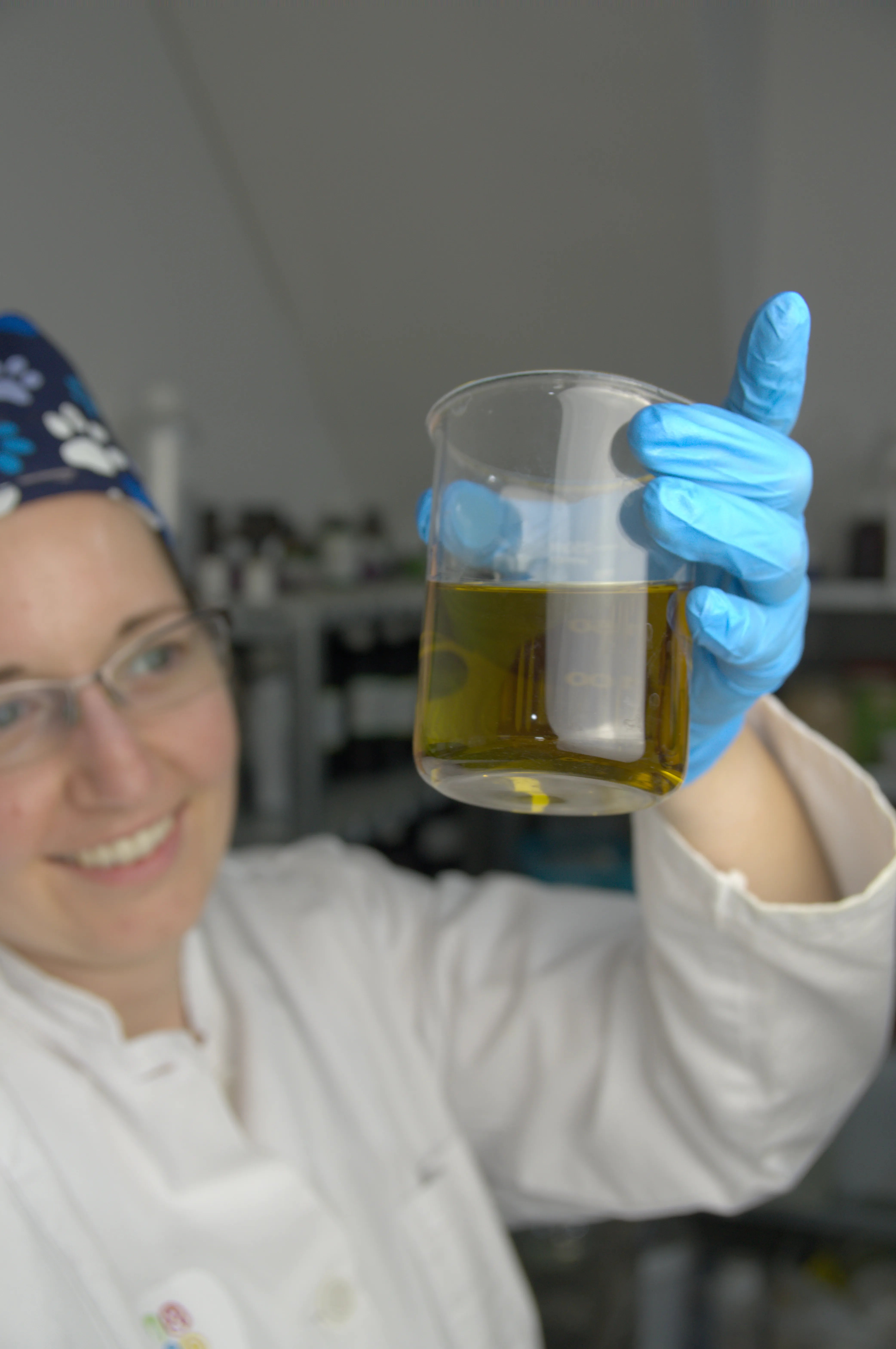 Eine Frau in weißem Kittel hält lächelnd ein Becherglas mit grünem Olivenöl in einem Labor hoch.