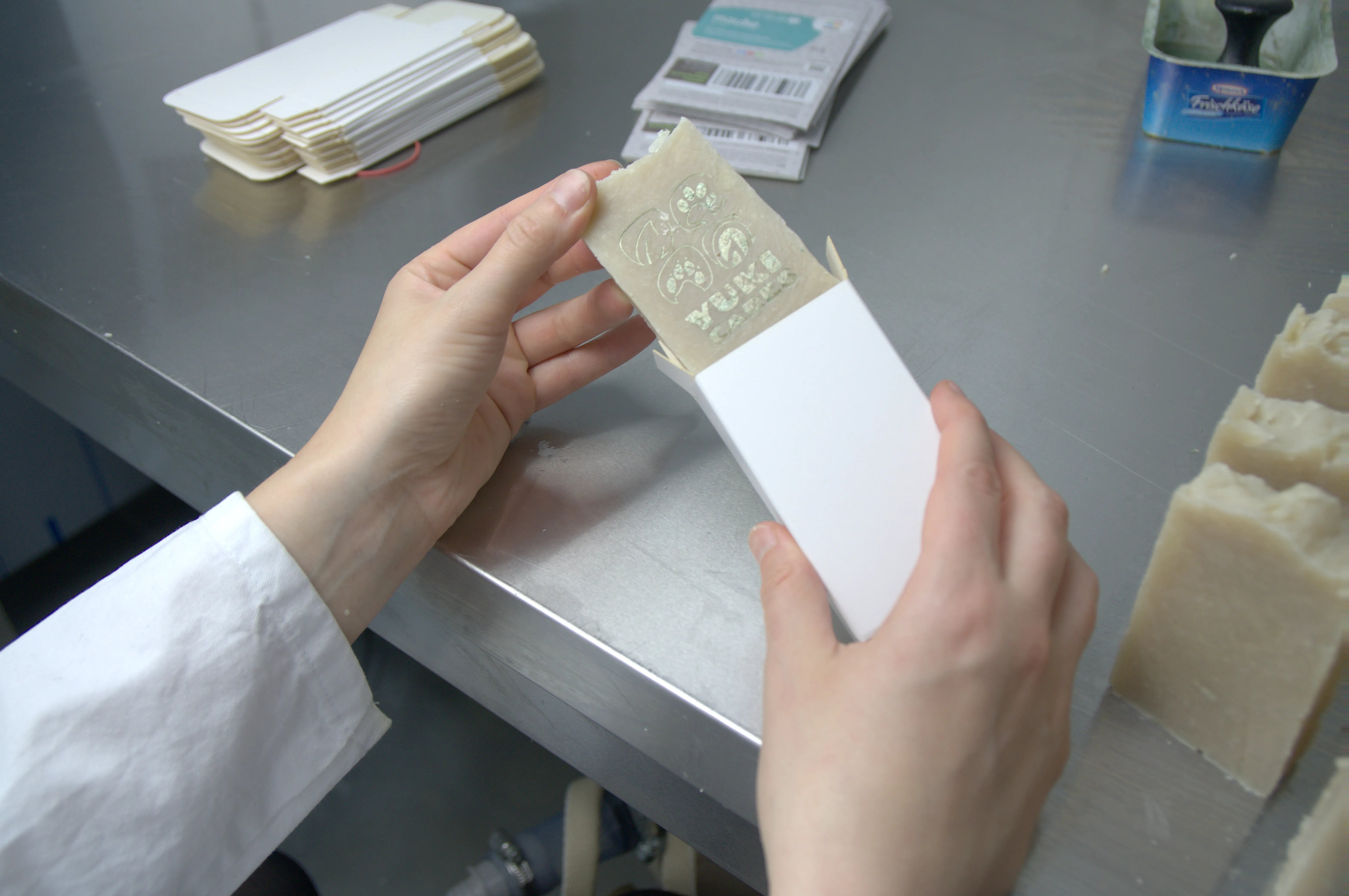 Eine Person öffnet ein weißes Pappetui und zeigt eine geprägte Seife auf einem Metalltisch.