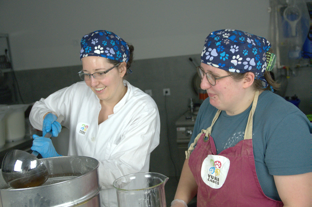 Zwei Frauen mit bunten Kopftüchern arbeiten lächelnd in einer Küche und schöpfen Kokosöl aus einem Fass.