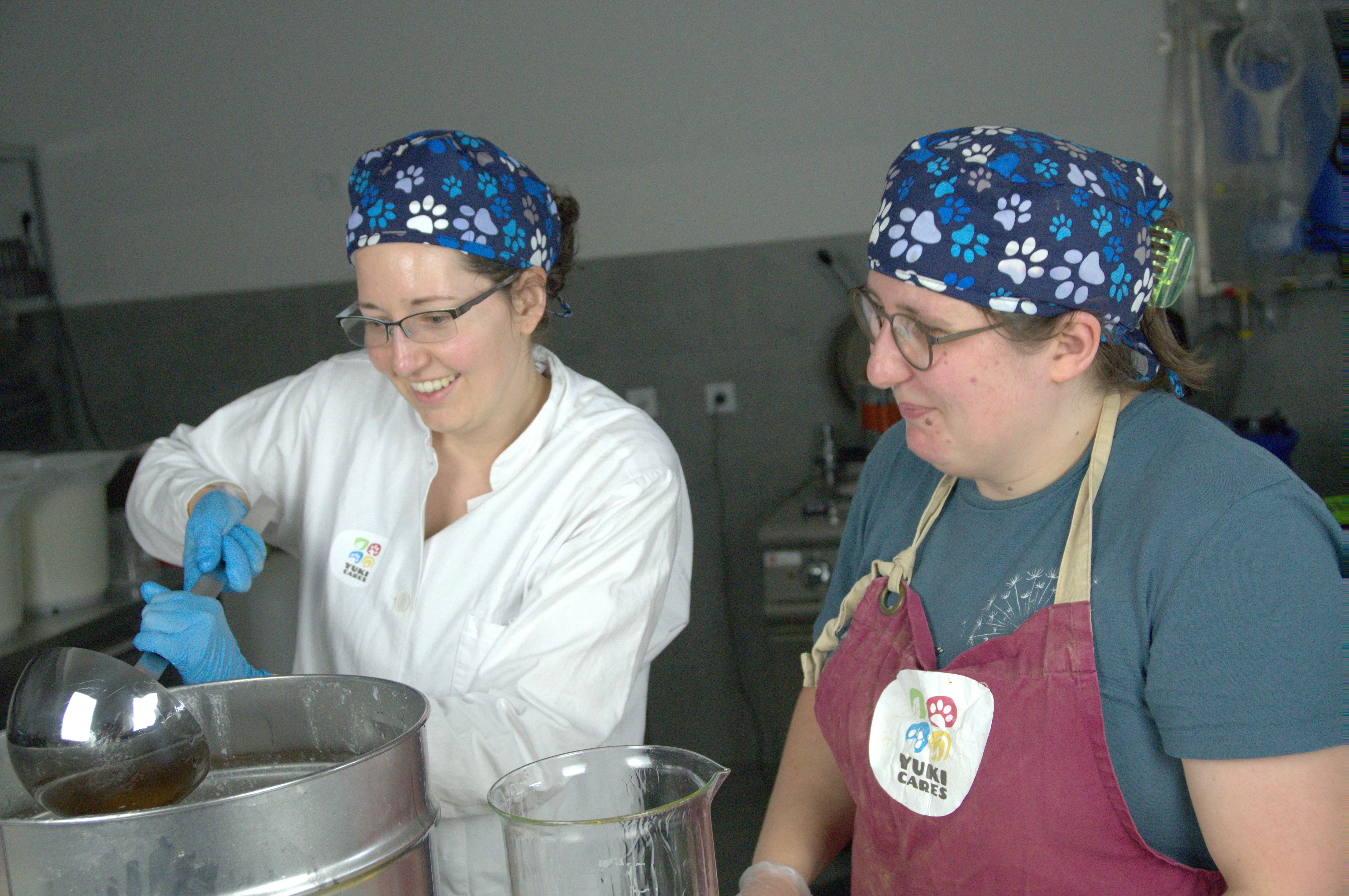 Zwei Frauen mit bunten Kopftüchern arbeiten lächelnd in einer Küche und schöpfen Kokosöl aus einem Fass.