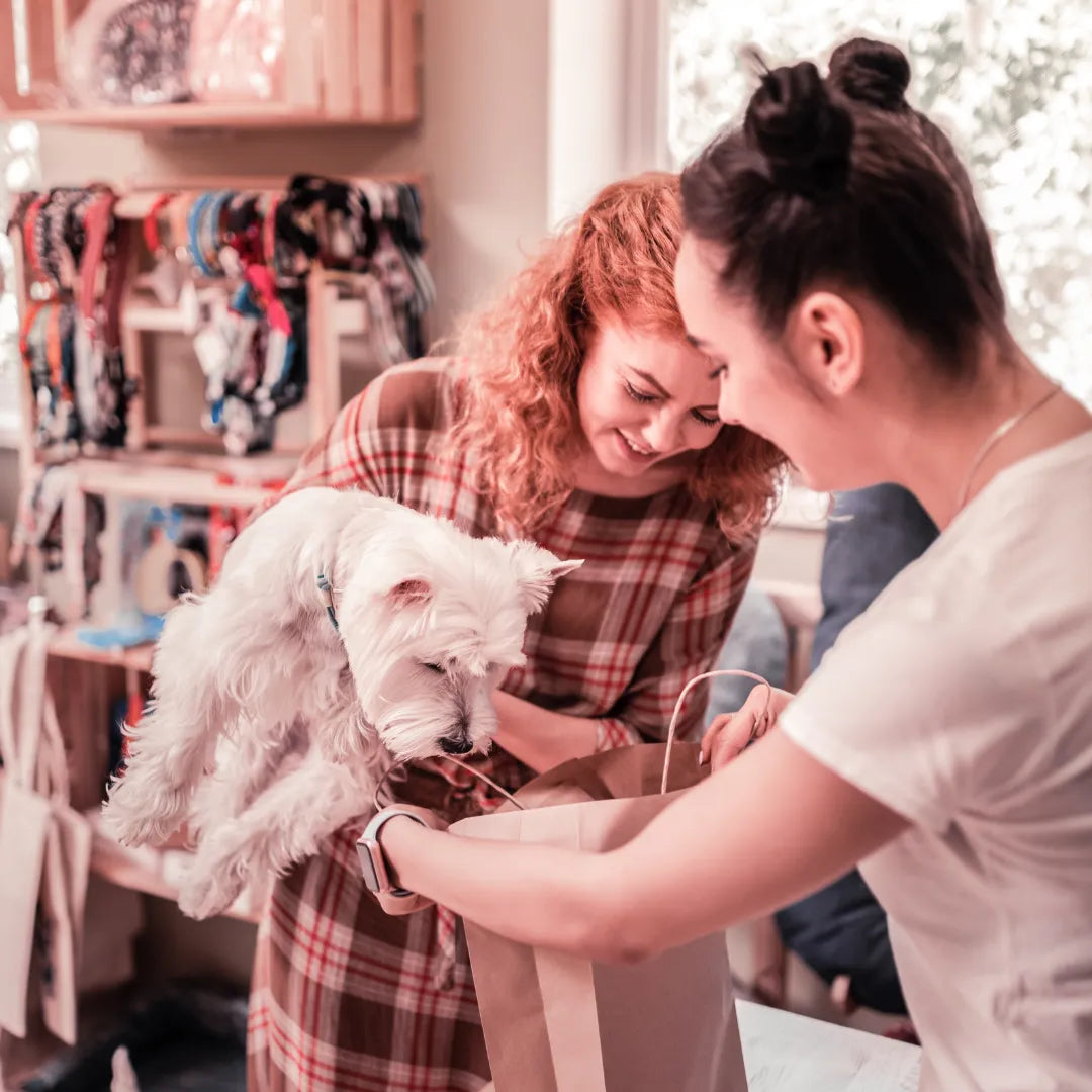 Zwei Frauen lächeln, während eine von ihnen einen kleinen weißen Hund hält und die andere in eine Einkaufstasche schaut.