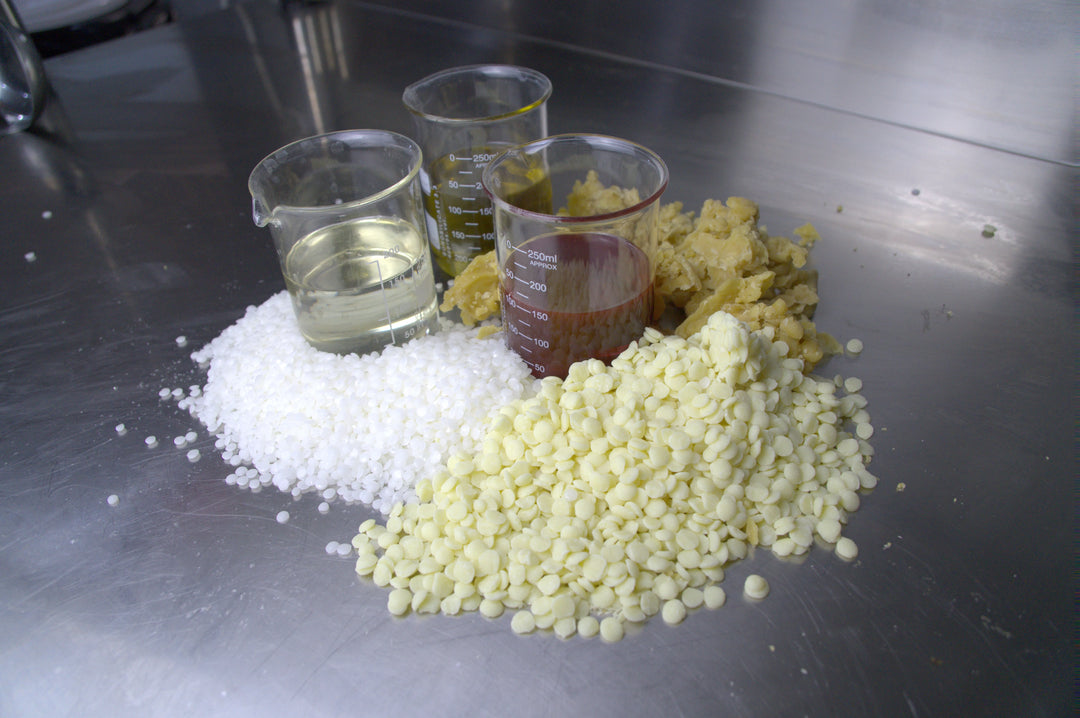 Drei Messbecher mit unterschiedlichen Flüssigkeiten stehen auf einem Tisch, umgeben von weißen Montanov und gelben Kakaobutterpellets, sowie Bienewachsbrocken.