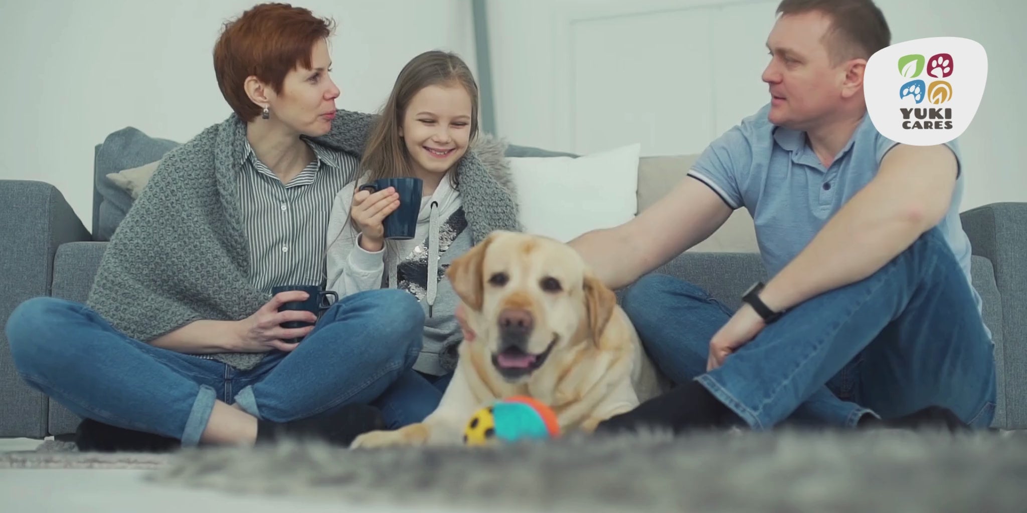 Eine Familie sitzt auf dem Teppich auf dem Boden, Vater, Mutter und kleine Tochter, ihr Hund sitzt in der Mitte und wird gestreichelt. Die Szene sieht sehr kuschelig aus und Mutte rund Tocher haben eetassen in der Hand.