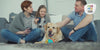 Eine Familie sitzt auf dem Teppich auf dem Boden, Vater, Mutter und kleine Tochter, ihr Hund sitzt in der Mitte und wird gestreichelt. Die Szene sieht sehr kuschelig aus und Mutte rund Tocher haben eetassen in der Hand.